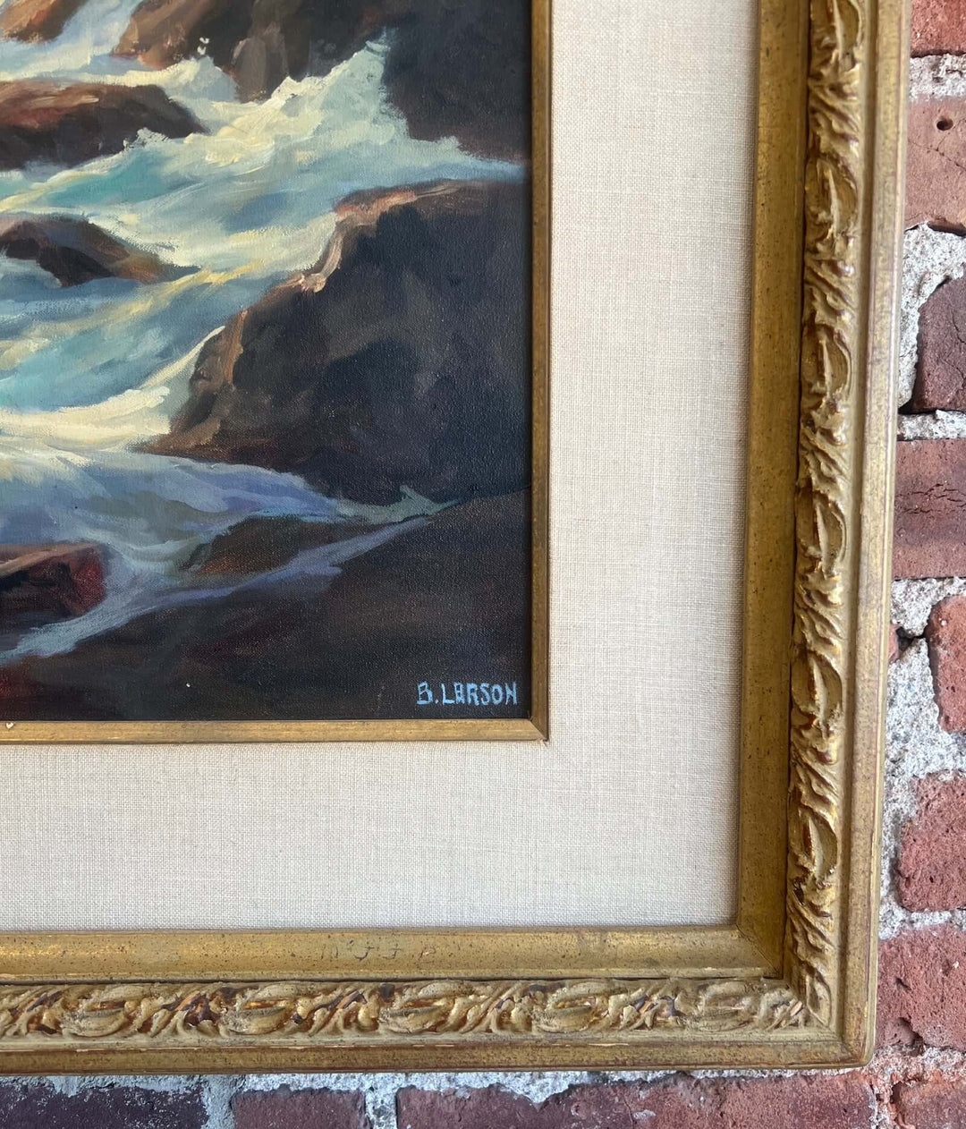Framed painting of a rocky shoreline with waves, signed B. Larson, on a textured wall.